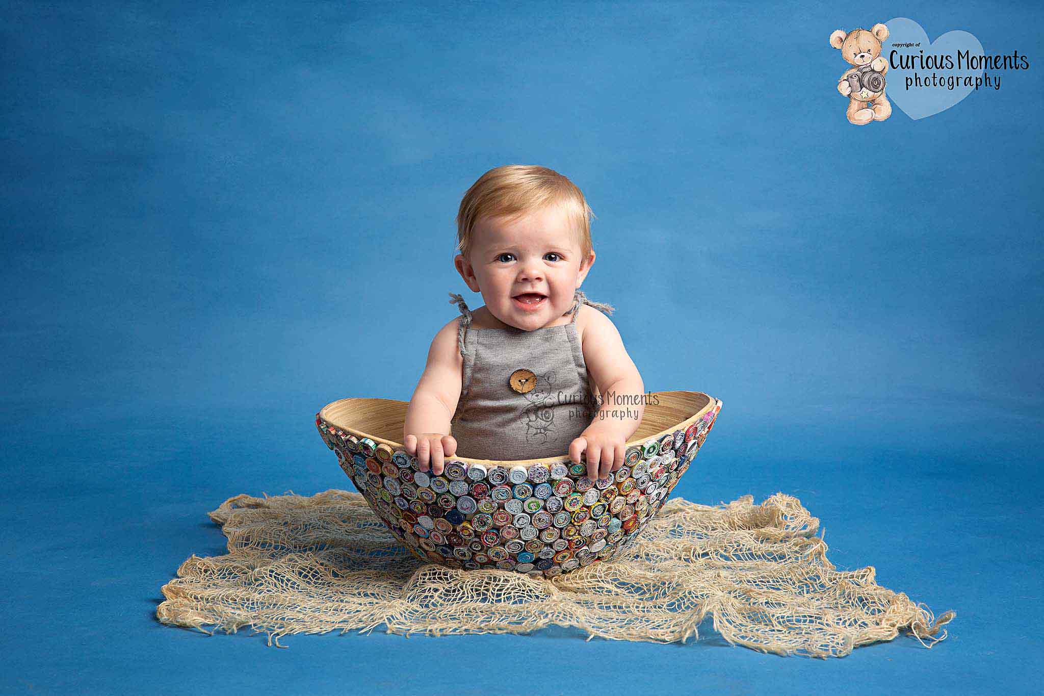 baby boy wearing a grwy romper sitting in a muticoloured bowl on a blue background