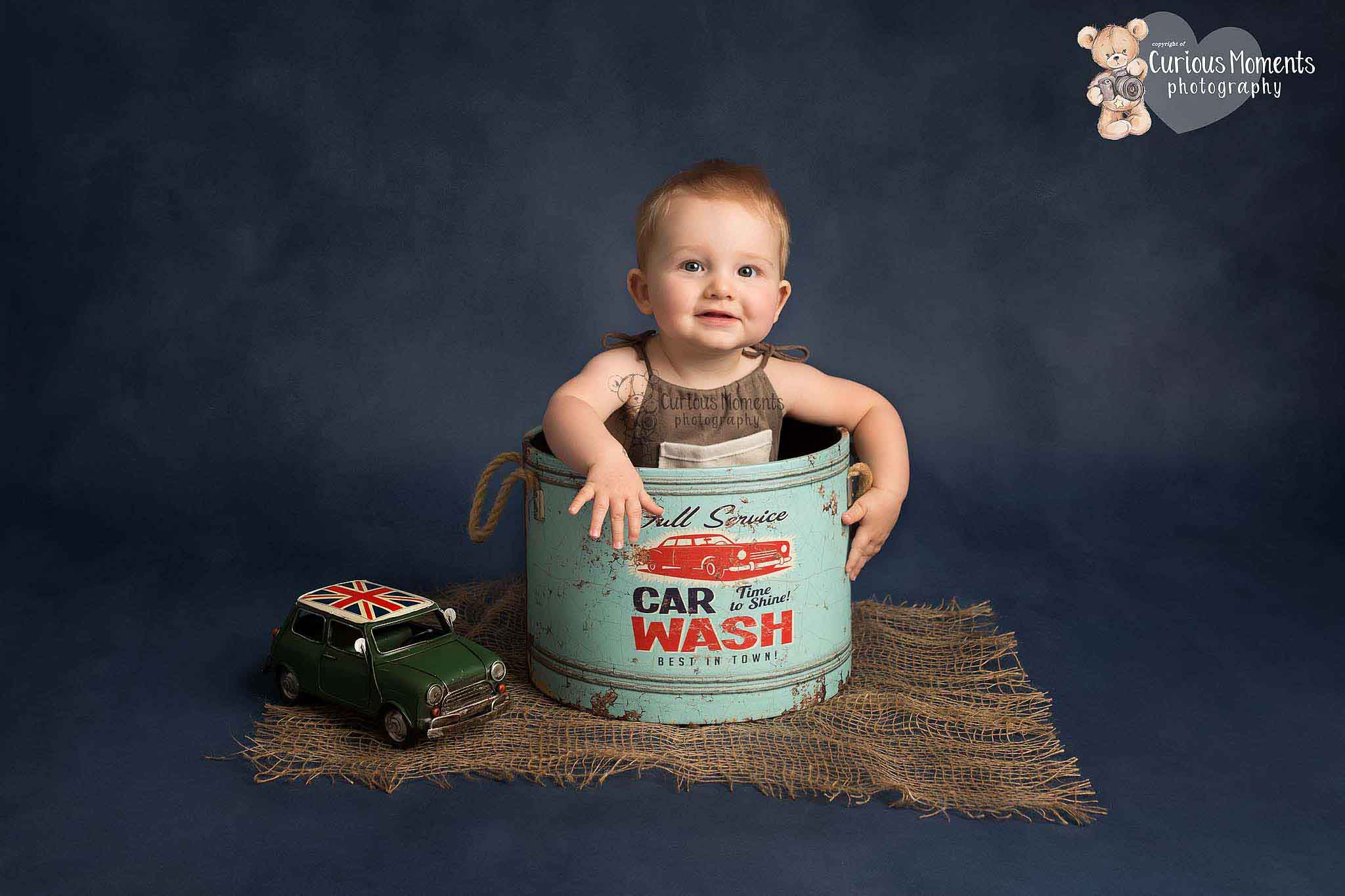 Baby boy sat in a car wash inspired tub next a mini car toy on a blue background