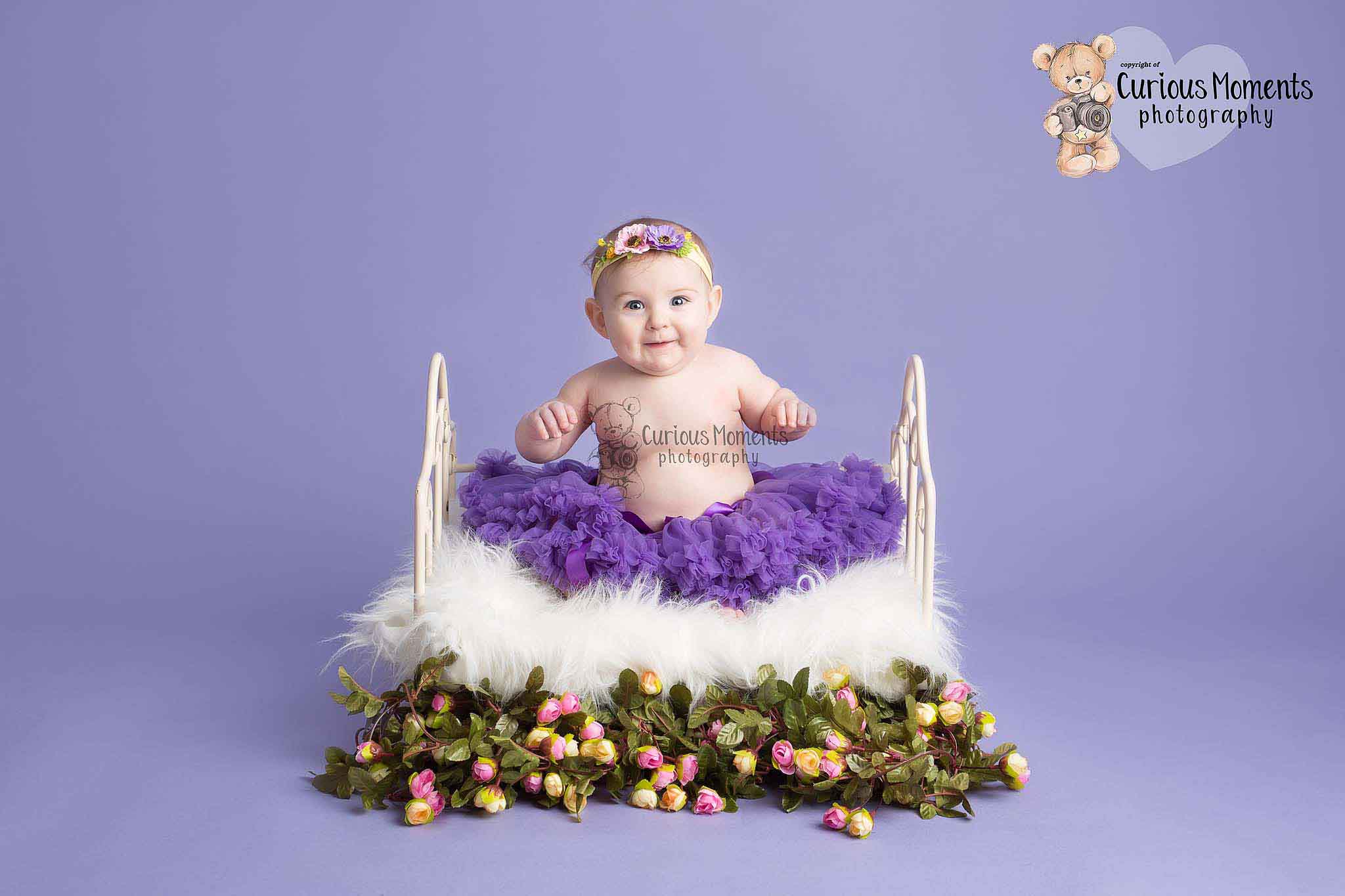 Happy baby firl wearing a purple tutu sat on a metal bed surrounded by flowers on a purple background during her photo shoot with pembrokeshire baby photographer
