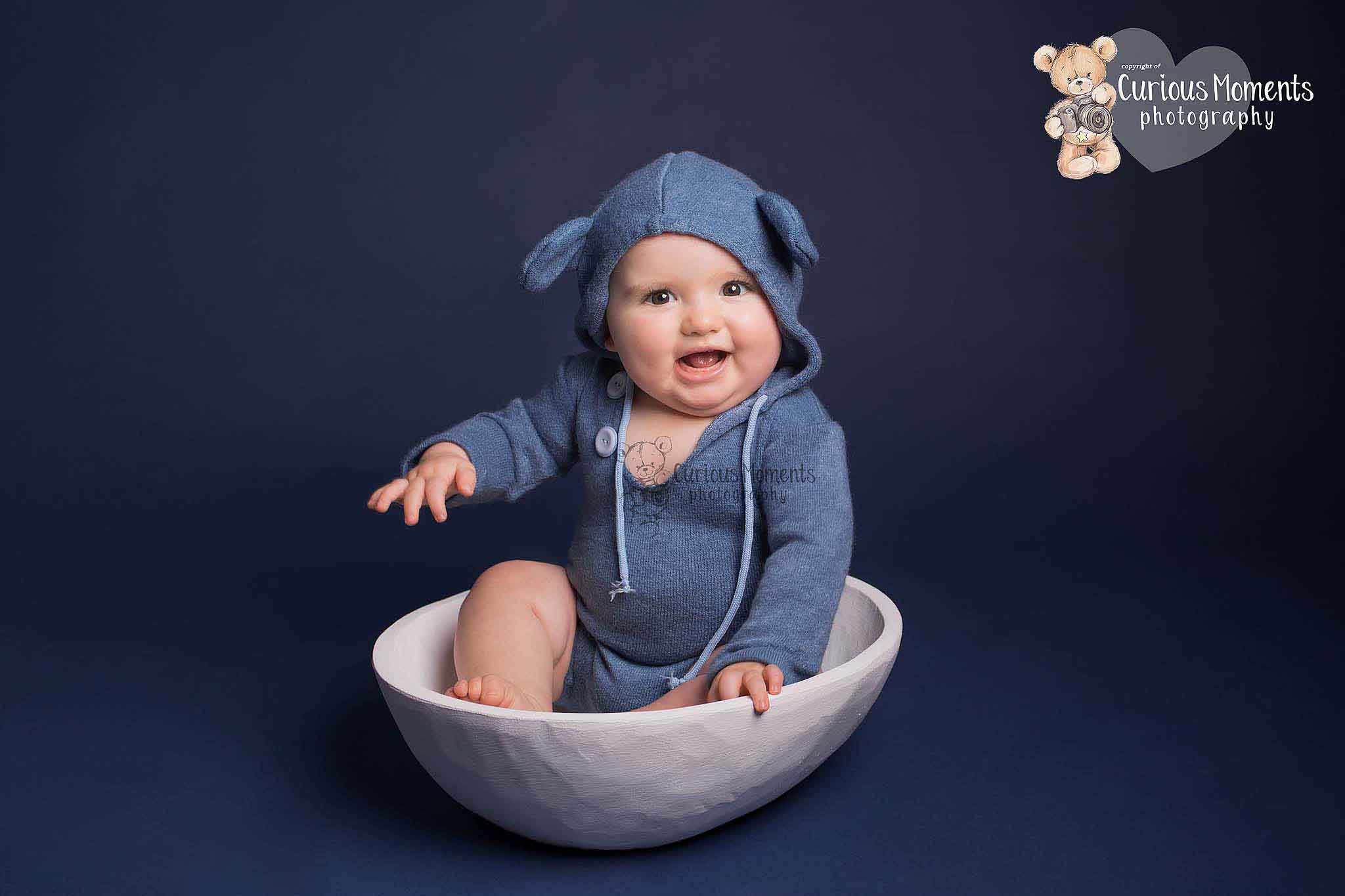 Happy smiling baby boy sat in a white bowl on a blue background wearing a blue bear style romper by Pembrokeshire baby photographer