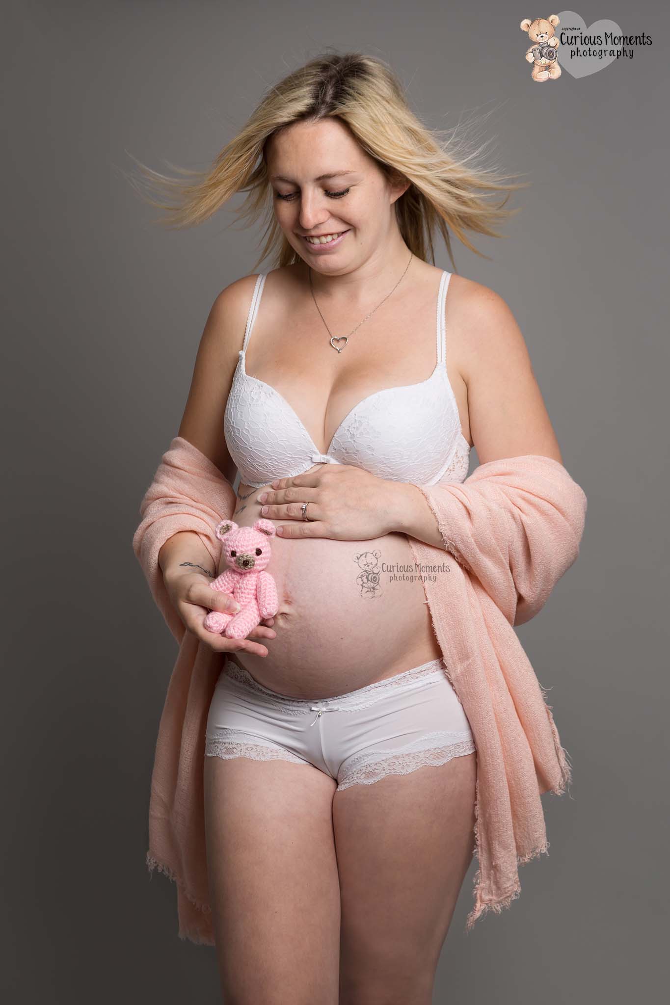 Pregnant lady expecting baby girl wearing white underwaer with off the shoulder pink drape holding pink teddy bear during her pregnancy photoshoot
