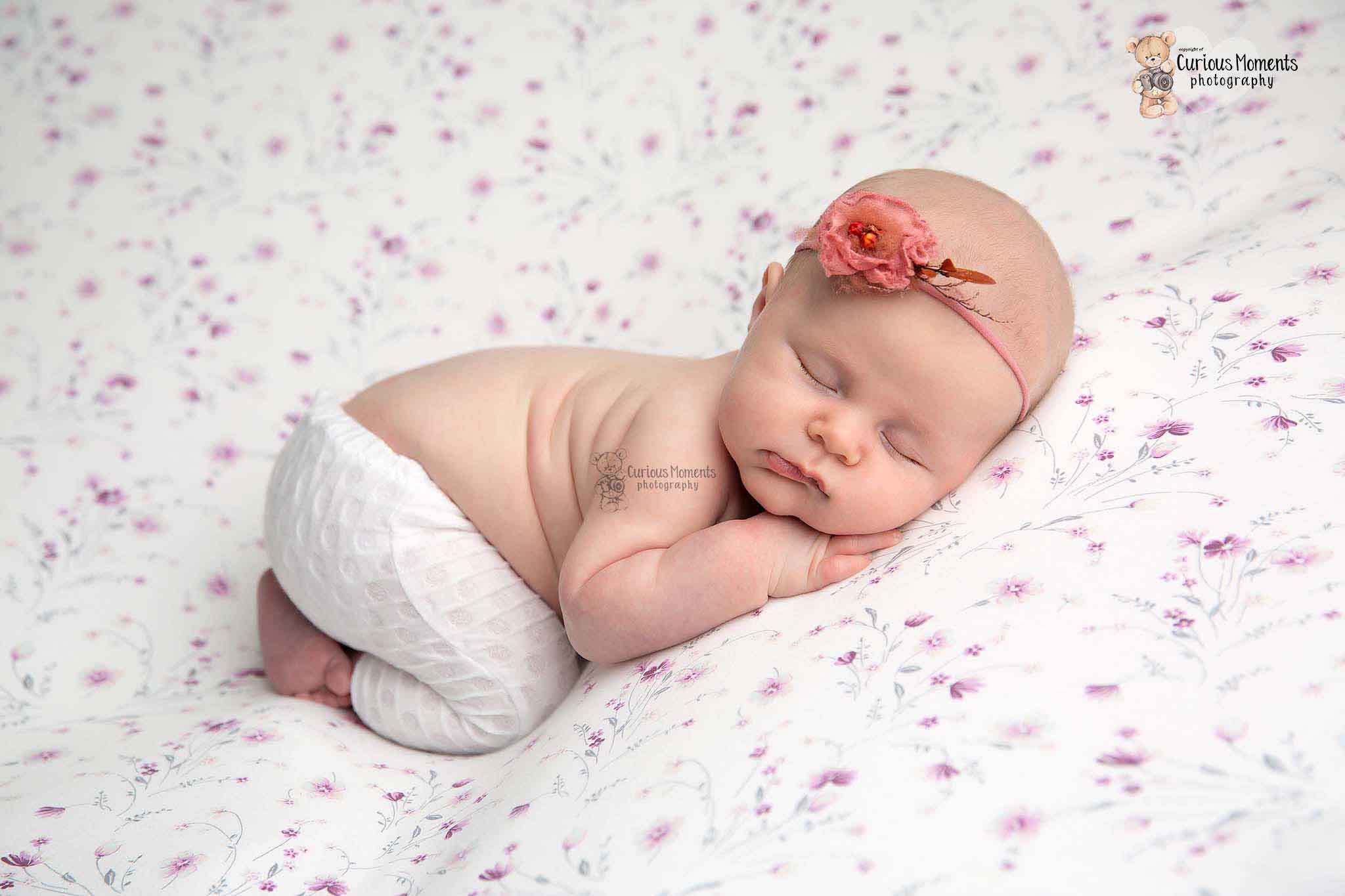 newborn baby girl in bum up pose wearing white trousers and pink hairband and on a floral background
