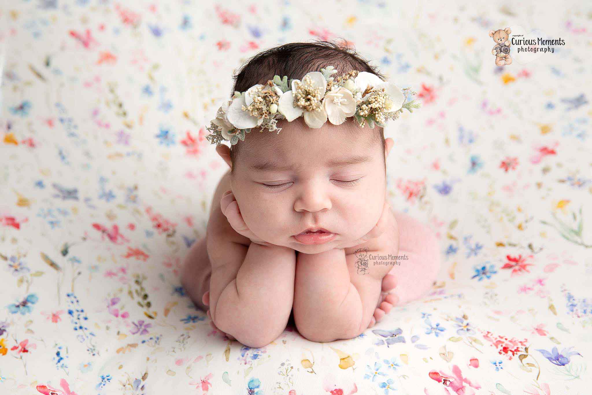newborn baby girl in froggy pose on a multicolour floral background and wearing a white floral crown