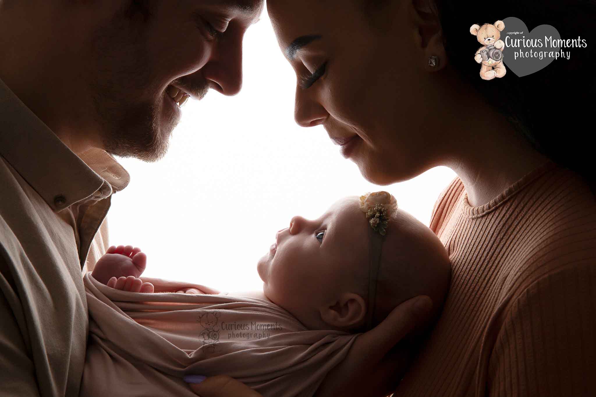 Backlit image of new parents looking down at their new baby girl by Curious moments photography